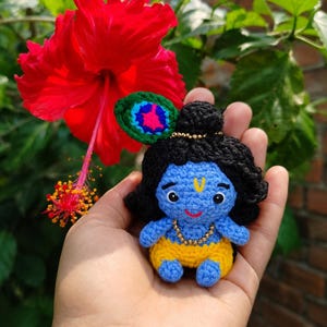 Puede incluir: Una pequeña muñeca tejida a ganchillo a mano que representa una figura de piel azul con cabello negro, vistiendo pantalones amarillos y un collar de cuentas doradas. La muñeca incluye un accesorio de pluma de pavo real verde, azul y rosa.