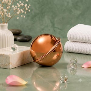 May include: A polished copper-colored spherical object with a thin ring encircling its center. The smooth, reflective metal sphere sits on a surface alongside small clear glass spheres, a white marble block, and pink flower petals.