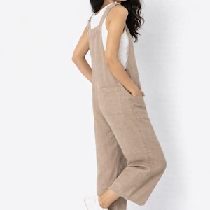May include: A tan linen jumpsuit with wide legs and a square neckline. The jumpsuit has two front pockets and is worn with a white tank top and white sneakers. The model is smiling and looking over their shoulder.