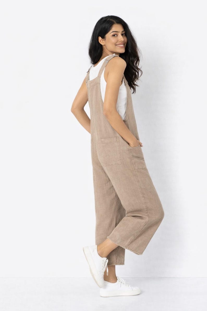 May include: A tan linen jumpsuit with wide legs and a square neckline. The jumpsuit has two front pockets and is worn with a white tank top and white sneakers. The model is smiling and looking over their shoulder.