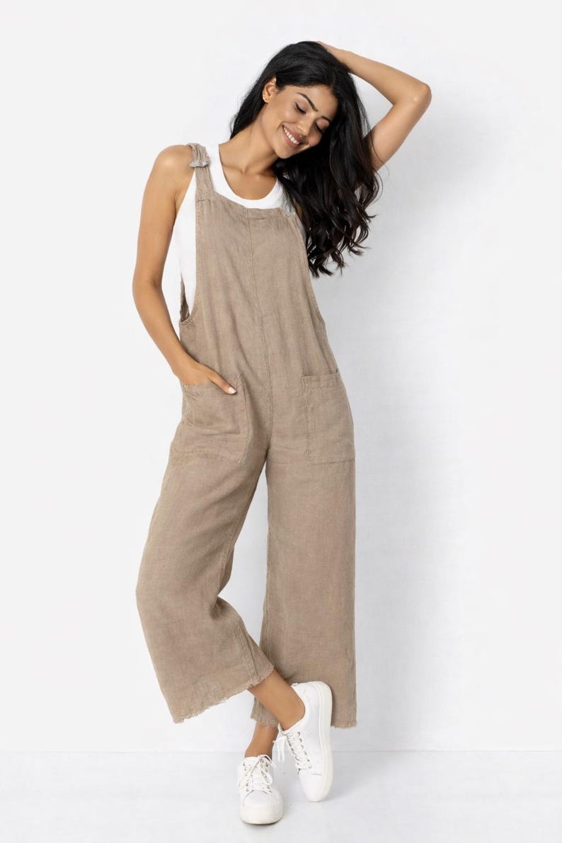 May include: A woman wearing a tan linen jumpsuit with a white tank top underneath. The jumpsuit has wide legs, front pockets, and adjustable straps. She is wearing white sneakers. The background is plain white.