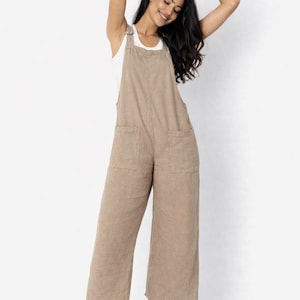 May include: A tan linen jumpsuit with wide legs and two front pockets. The jumpsuit has a bib-style top with adjustable straps. The model is wearing a white tank top underneath and white sneakers.