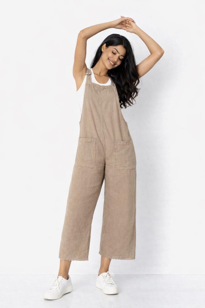 May include: A tan linen jumpsuit with wide legs and two front pockets. The jumpsuit has a bib-style top with adjustable straps. The model is wearing a white tank top underneath and white sneakers.