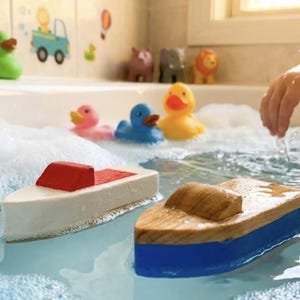 May include: A child's bath scene featuring toy boats and rubber ducks. Two wooden boats with red and brown details float in the water. Several colourful rubber ducks and animal figurines are visible. Bubbles fill the water, and a child's hand reaches in.
