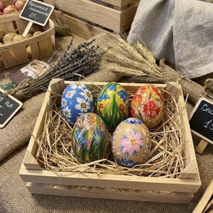 Puede incluir: Una caja de madera llena de huevos de Pascua decorativos. Los huevos están pintados a mano con diseños florales en varios colores, incluyendo azul, amarillo, rojo y rosa. La caja está sobre una superficie de arpillera.