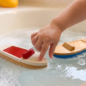 May include: Two wooden toy boats floating in water with soap bubbles. One boat features a natural wood hull with a red deck and a small red cabin. The second boat has a natural wood deck and a blue hull. Both boats have a simple, smooth design.