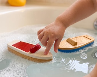 May include: Two wooden toy boats floating in water with soap bubbles. One boat features a natural wood hull with a red deck and a small red cabin. The second boat has a natural wood deck and a blue hull. Both boats have a simple, smooth design.