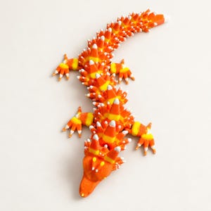 May include: A lizard-shaped articulated figure constructed from numerous candy corn pieces. The creature features a bright orange head and limbs, with a body covered in spikes coloured in horizontal bands of orange, yellow, and white.