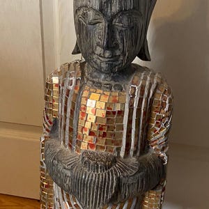 May include: A wooden Buddha statue with a dark finish, adorned with mosaic tiles in gold, red, and silver. The statue is on a black base and features a serene expression.