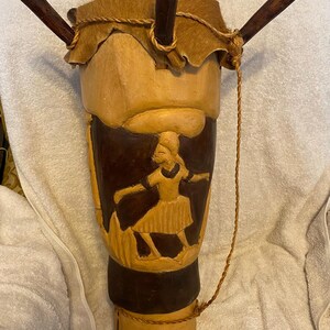 May include: Hand-carved wooden drum with a natural finish and a dark brown base. The drum features a carved relief of a person carrying something on their head. The drumhead is made of animal hide and secured with a rope. Three wooden sticks are attached to the top.