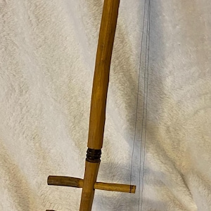 May include: A handmade stringed musical instrument with a rounded, gourd-like body and a long wooden neck. The instrument has three horizontal pegs and black strings. The body is tan with a dark geometric design.