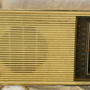 May include: Vintage Sylvania radio with a gold-toned, horizontally-lined front panel. The right side features a black panel with a tuning dial, frequency numbers, and orange knobs. The Sylvania logo is visible on the lower left.