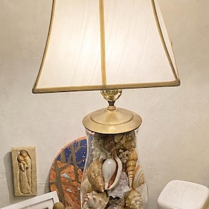 May include: A decorative table lamp with a glass base filled with seashells. The lamp has a brass-colored base and a rectangular lampshade. The lamp is surrounded by decorative figurines and artwork.