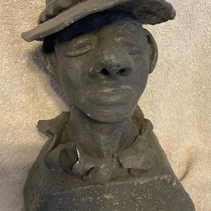 May include: A dark grey bust sculpture of a person wearing a hat. The sculpture has a textured surface and detailed facial features, including closed eyes and a prominent nose. The hat has a wide brim.