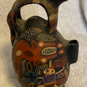 May include: A black ceramic vessel with a monkey-like figure on top and a handle. The vessel is decorated with colorful abstract patterns in red, white, and yellow. The base is black and the vessel has a spout.