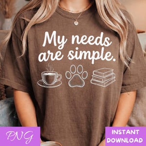 May include: Brown t-shirt with the text "My needs are simple." Below the text are white illustrations of a coffee cup, a paw print, and a stack of books. The shirt has a relaxed fit.