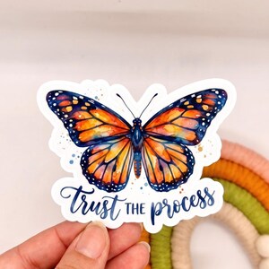 Può includere: Un adesivo fustellato raffigurante una farfalla monarca in stile acquerello con ali arancioni vivaci e blu scuro. Sotto la farfalla, il testo "Trust the process" è scritto in blu scuro con un'elegante grafia corsiva su sfondo bianco.