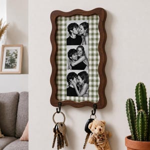 May include: A rectangular wall-mounted key holder with a wavy dark wood frame and a green and white gingham background. It features a vertical strip of three black and white photographs of a couple and two black metal hooks holding key rings.