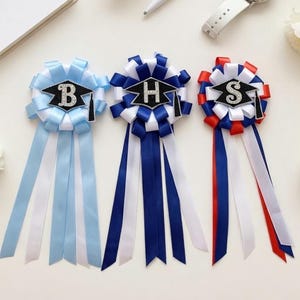 May include: Three graduation rosettes with ribbons in blue, navy, and red. Each rosette has a letter and a graduation cap design. A white watch, pen, and notebook are also in the image.