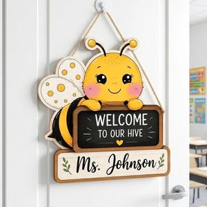May include: A decorative wooden wall sign shaped like a cartoon bee with yellow and black stripes, holding a black chalkboard sign that reads WELCOME TO OUR HIVE with a yellow heart, and a lower white sign with the text Ms. Johnson in black script.