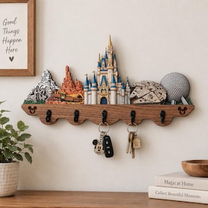 Può includere: Un portachiavi da parete in legno con cinque ganci in metallo nero e una decorazione superiore che raffigura strutture iconiche di parchi a tema, tra cui un castello, una montagna, un paesaggio desertico, un'astronave e una cupola geodetica.