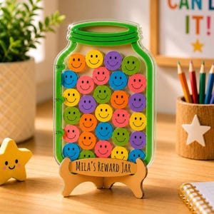May include: A clear acrylic jar-shaped reward container with a green border, filled with colorful wooden tokens featuring smiley faces. The base is a wooden stand engraved with the text Mila's Reward Jar. The jar includes markings for 25%, 50%, and 75% capacity levels.