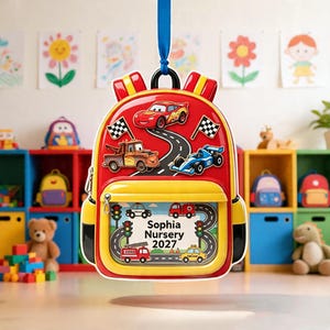 May include: A red and yellow backpack ornament featuring Lightning McQueen, a tow truck, and a race car on a track. The front pocket has a clear window displaying a road scene with traffic lights, vehicles, and the text Sophia Nursery 2027.