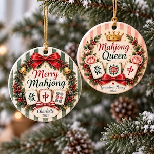 Può includere: Due ornamenti rotondi in ceramica con sfondi a righe verdi e bianche, decorati con ghirlande di pino, bacche rosse e fiocchi rossi. Uno riporta Merry Mahjong con Charlotte 2026, l'altro Mahjong Queen con Grandma Nancy 2026.
