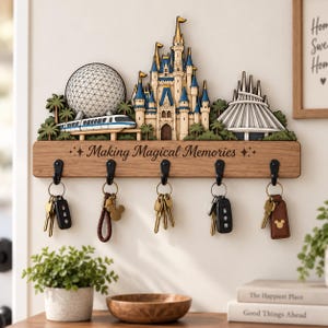 Könnte beinhalten: Ein hölzernes Wand-Schlüsselbrett mit einem mehrschichtigen Ausschnitt-Design eines Themenpark-Schlosses, einer geodätischen Kuppel und eines futuristischen Berges. Das Brett ist mit Making Magical Memories graviert und hat fünf schwarze Metallhaken.