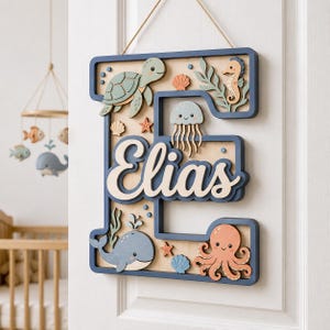 Könnte beinhalten: Ein hölzernes Wandobjekt in Form des Buchstabens E mit dem Namen Elias in weißer Schreibschrift. Das blau umrandete Schild ist mit geschichteten Ausschnitten einer Meeresschildkröte, eines Seepferdchens, einer Qualle, eines Wals und Muscheln verziert.