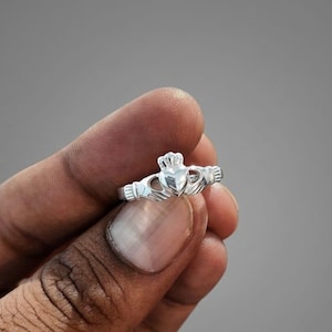 Könnte beinhalten: Ein polierter Claddagh-Ring aus Silber mit einem zentralen Herz, das von einer Krone gekrönt und von zwei Händen flankiert wird. Der Ring besteht aus glänzendem Metall und zeigt die traditionellen Gestaltungselemente aus Herz, Händen und Krone.