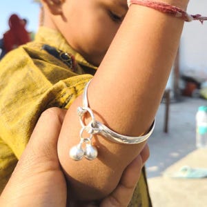 Könnte beinhalten: Ein polierter Silberarmreif mit einem glatten, geschwungenen Band und zwei kleinen, runden Glöckchen, die an einer zentralen Schlaufe befestigt sind. Das metallische Accessoire wird am Oberarm getragen und hat eine glänzende Oberfläche.