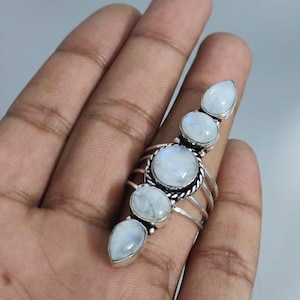 Könnte beinhalten: Ein Silberring mit fünf Mondstein-Cabochons, die in einer vertikalen Linie angeordnet sind. Die Steine haben unterschiedliche Formen, darunter runde und tropfenförmige Schliffe, gefasst in poliertem Silbermetall. Die Ringschiene besteht aus mehreren.