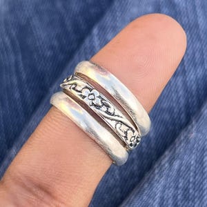 Könnte beinhalten: Ein Stapel von drei silbernen Ringen. Der mittlere Ring hat ein Blumenmuster. Die Ringe werden an einem Finger getragen, vor einem blauen Jeanshintergrund. Die Ringe haben eine Vintage- oder Boho-Ästhetik.