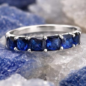 Könnte beinhalten: Ein silberner Ring mit einer Reihe quadratisch geschliffener, tiefblauer Edelsteine. Der Ring ist vor einem Hintergrund aus blauem und weißem strukturiertem Stein platziert, der die lebendige Farbe der Edelsteine hervorhebt.
