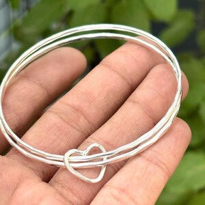 Könnte beinhalten: Zwei silberne Armreifen mit einem herzförmigen Anhänger. Die Armreifen bestehen aus glänzendem, strukturiertem Metall und werden in einer Hand vor einem verschwommenen grünen Hintergrund gehalten. Der Herzanhänger ist mittig auf den Armreifen angebracht.