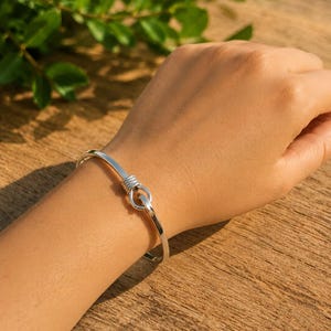 Könnte beinhalten: Ein silberfarbenes Armreif-Armband mit Haken- und Ösenverschluss. Das Armband hat ein schlichtes, elegantes Design mit glatter, polierter Oberfläche. Der Haken- und Ösenverschluss ist ein dekoratives Element, das für visuelles Interesse sorgt. Das Armband wird am Handgelenk getragen.