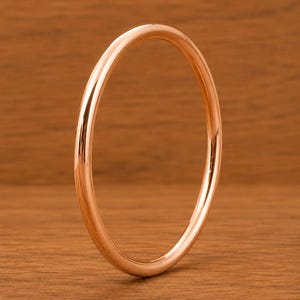 Könnte beinhalten: Ein roségoldenes Armreif-Armband. Das kreisförmige Armband ist glatt und poliert und reflektiert das Licht. Der Schmuck wird auf einer Holzoberfläche präsentiert.