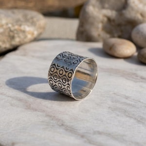 Könnte beinhalten: Ein breiter Silberring mit strukturierter Oberfläche. Der Ring hat ein sich wiederholendes Muster aus schwarzen und goldenen Kreisformen. Der Ring wird auf einer Marmoroberfläche mit Felsen im Hintergrund präsentiert. Schmuck.