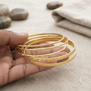 Könnte beinhalten: Ein Set aus fünf goldfarbenen Metallarmreifen mit unterschiedlichen Texturen und Mustern. Die Kollektion umfasst ein Perlenband, ein glattes Wellenband und drei Bänder mit komplexen floralen und geometrischen Gravuren, die zusammen gestapelt sind.