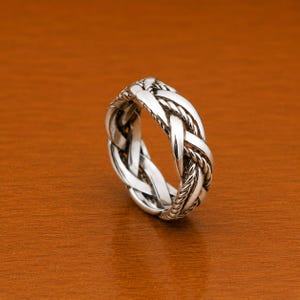 Könnte beinhalten: Ein silberfarbener Ring mit geflochtenem Design. Der Ring hat ein gewebtes Muster mit mehreren Strängen, wodurch ein strukturiertes und kompliziertes Aussehen entsteht. Der Ring ist vor einem warmen, braunen Hintergrund platziert.