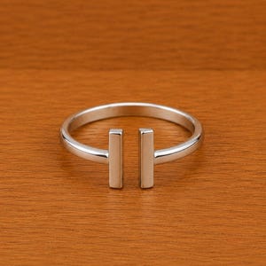 Könnte beinhalten: Ein silberfarbener Ring mit minimalistischem Design. Der Ring hat ein Band mit zwei vertikalen rechteckigen Stäben. Der Ring wird auf einer Holzoberfläche präsentiert. Der Ring ist ein Schmuckstück.