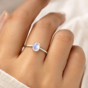 Könnte beinhalten: Ein silberner Ring mit einem ovalen, schillernden Mondstein. Der Mondstein ist in einer einfachen Vier-Krappen-Fassung gefasst. Der Ring wird an einem Finger gezeigt.