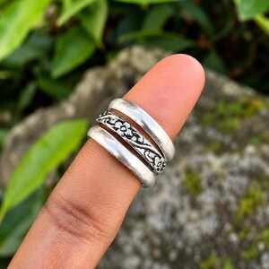Könnte beinhalten: Ein Set aus drei silberfarbenen Ringen, die übereinander getragen werden. Der obere und der untere Ring haben eine glatte, polierte Oberfläche, während der mittlere Ring ein geprägtes Blumenrankenmuster mit dunklen Vertiefungen aufweist.