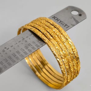 Könnte beinhalten: Ein Set aus vier goldfarbenen Armreifen mit strukturiertem Blumenmuster, platziert auf einem Metalllineal mit der Aufschrift KHYATI. Das Lineal zeigt, dass die Armreifen einen Innendurchmesser von etwa 6,35 cm haben, laut den Markierungen.