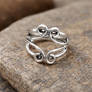 Könnte beinhalten: Ein silberfarbener Ring mit einem komplizierten, wirbelnden Design. Der Ring hat mehrere Schichten und dekorative, scrollartige Elemente, die einen einzigartigen und künstlerischen Look erzeugen. Der Ring ist vor einem neutralen, strukturierten Hintergrund platziert.