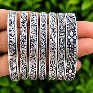 Könnte beinhalten: Sieben silberfarbene Metallarmreifen mit verschiedenen geprägten Mustern wie Elefanten, Blumen, Fischen, Om-Symbolen und geometrischen Formen. Jedes Armband hat ein dunkel oxidiertes Finish in den Vertiefungen, um die erhabenen Designs hervorzuheben.
