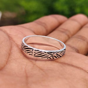 Könnte beinhalten: Ein silberner Ring mit einem detaillierten schwarzen Muster. Der Ring ist rund und hat ein strukturiertes Design um das Band. Der Ring wird auf einer Hand gezeigt.