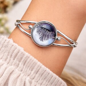 Könnte beinhalten: Silberfarbenes Armreif-Armband mit einem runden Anhänger mit einem dendritischen Muster in Lila und Weiß. Das Armband hat ein zartes, offenes Design mit kleinen dekorativen Kugeln. Ein stilvolles Schmuckstück.