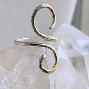 Könnte beinhalten: Ein silberfarbener Metallring mit offenem Bypass-Design und zwei ausgeprägten, spitz zulaufenden Spiralenden, die in entgegengesetzte Richtungen gebogen sind. Der Ring hat eine glatte, polierte Oberfläche und eine fließende S-förmige Gestalt.